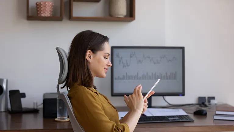 Confident analyst woman using app on smartphone, sitting at desktop monitor with trading infographics, sustainability charts, rates, chatting online, getting income, profit, smiling