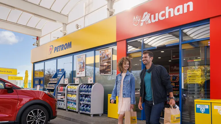 A woman and a man exit a Petrom Filling Station smiling. The visible branding includes the Petrom and MyAuchan logos.