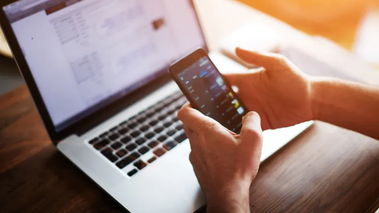 a man checks his phone while a laptop is seen in the background, in soft sun light